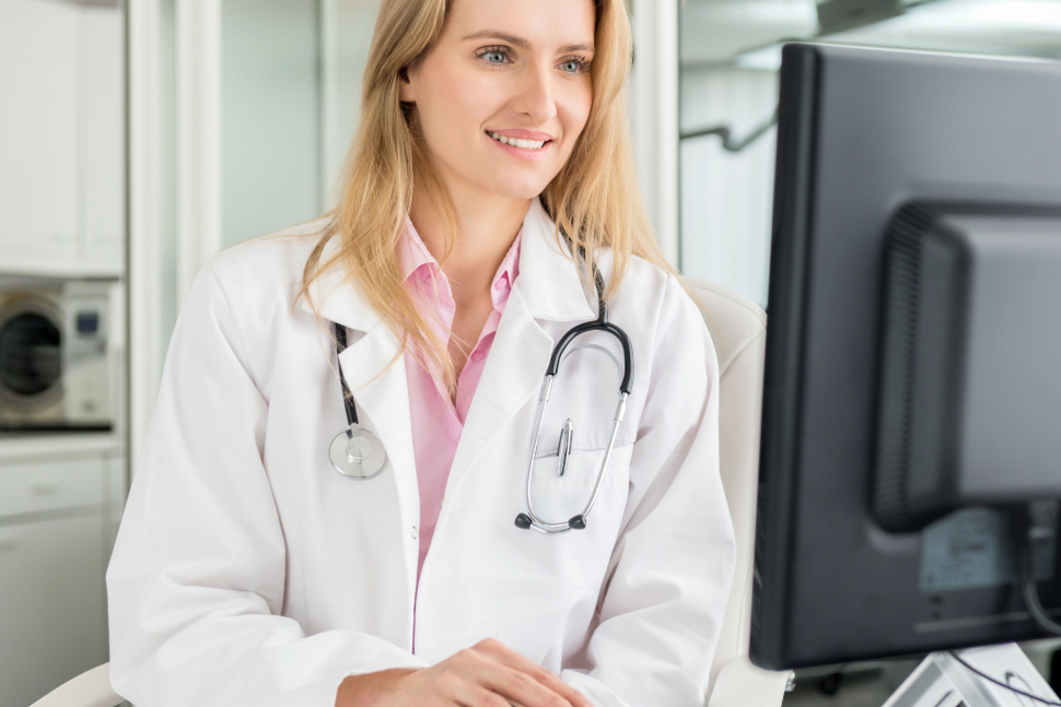 young smiling female doctor sitting at the desk and working on computer at hospital office